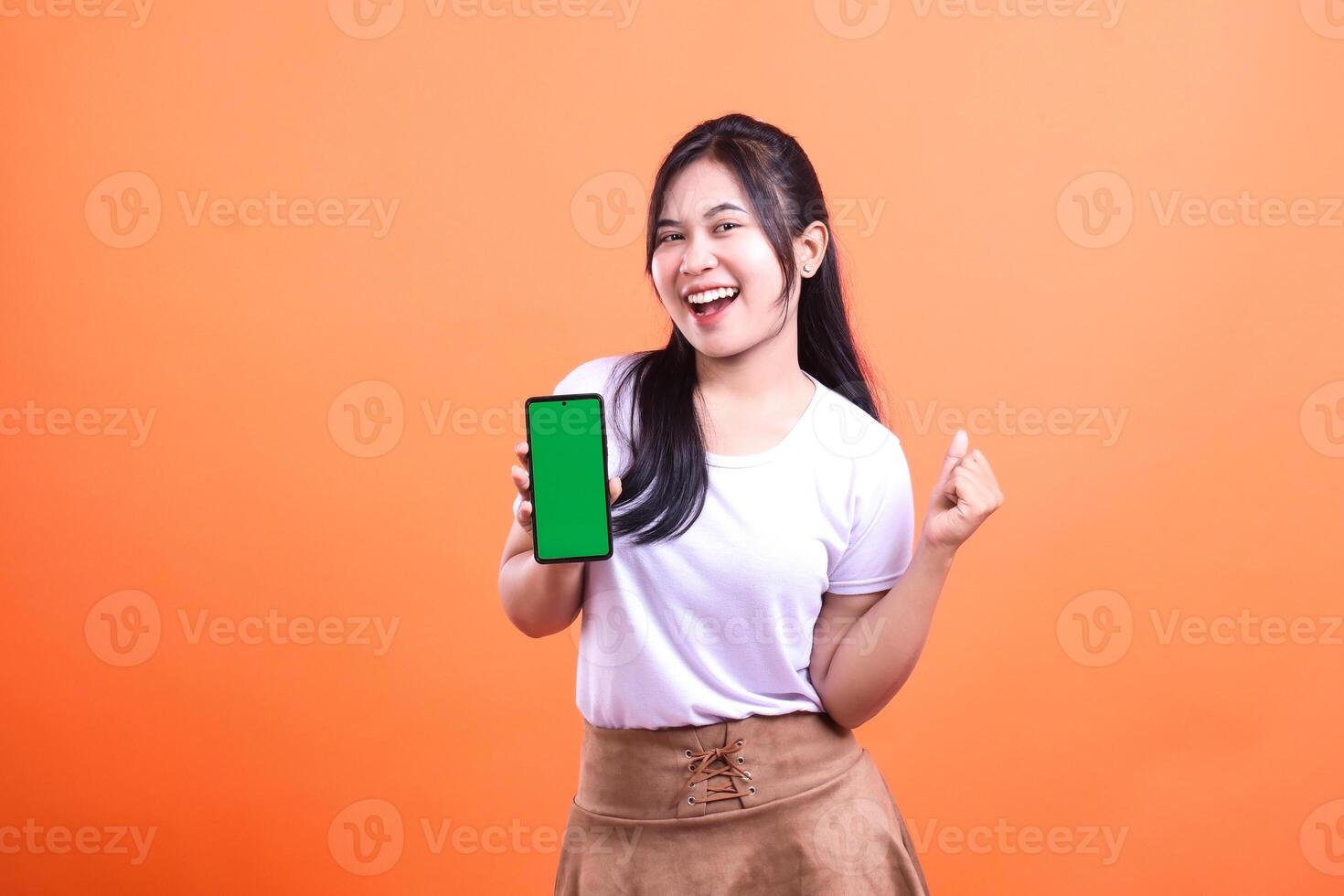 Woman showing smartphone with green screen against orange background smiling photo