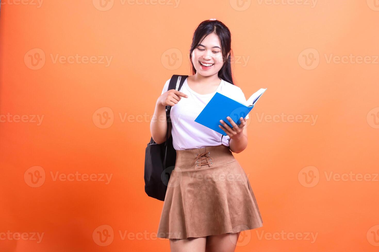 Cheerful student with a backpack reading a book, wearing a skirt and a white top and isolated on orange background photo