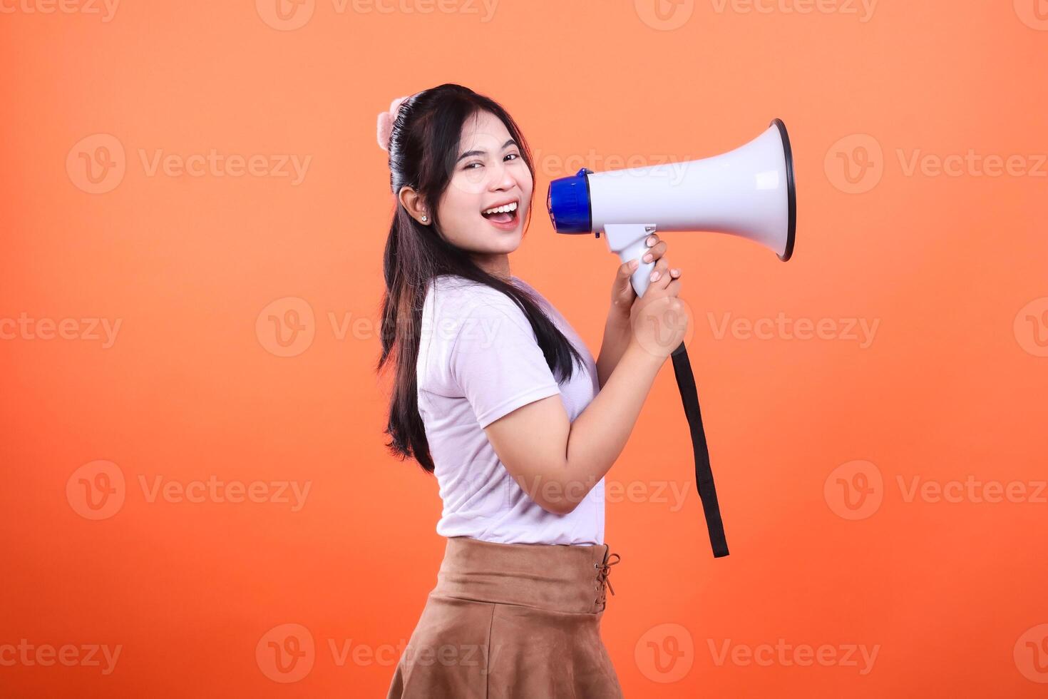 A woman asia with black hair and wearing a white shirt and brown skirt is holding a white megaphone and speaking into it. She is smiling while looking at the camera. isolated on an orange background. photo