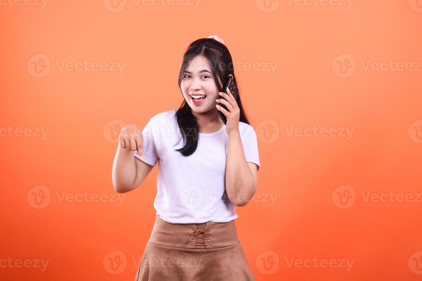Cheerful woman with phone pointing down, isolated on orange background, having a great time photo