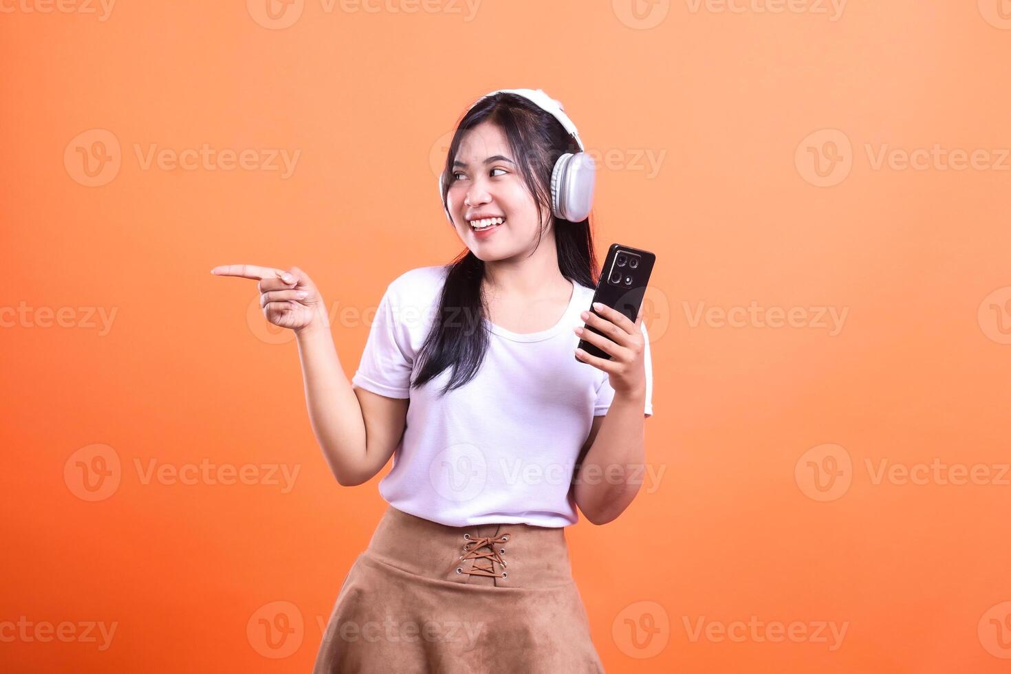 Smiling woman with headphones holding phone pointing to the left on orange backdrop photo