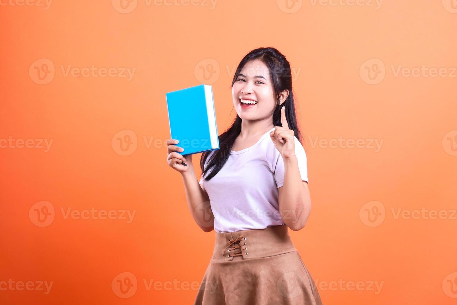 A woman holds a light blue book, with her right hand up and pointing, on an orange background photo