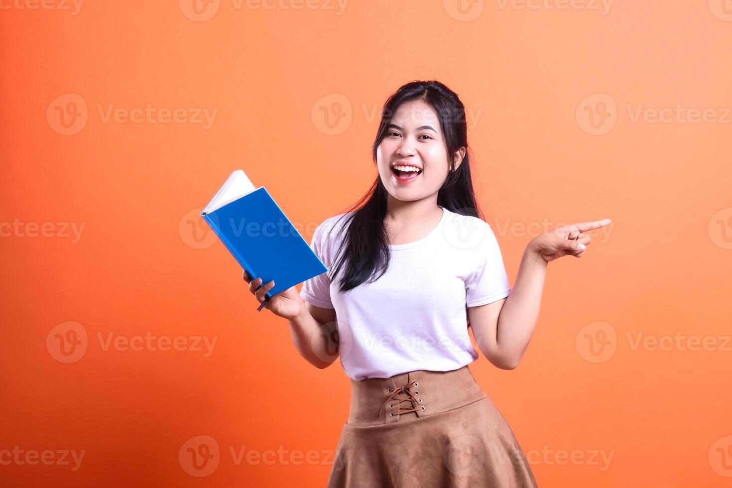 A woman holding a book and pointing with her right hand with a vibrant orange background color behind her photo