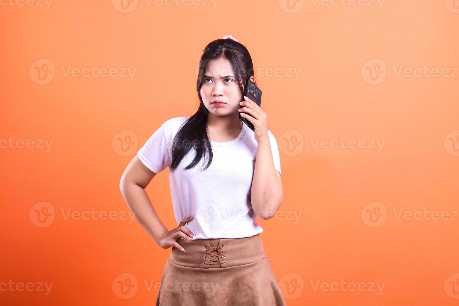 Woman with dark hair wearing a white shirt and a brown skirt talking on her phone on orange background photo