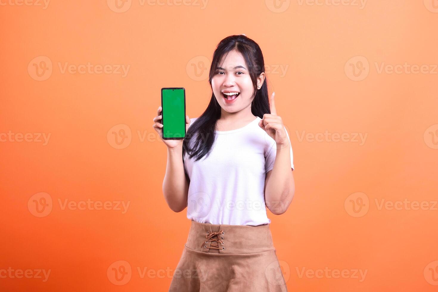 A woman with long black hair is holding a smart phone device with a green screen and raising a finger on an orange background. photo