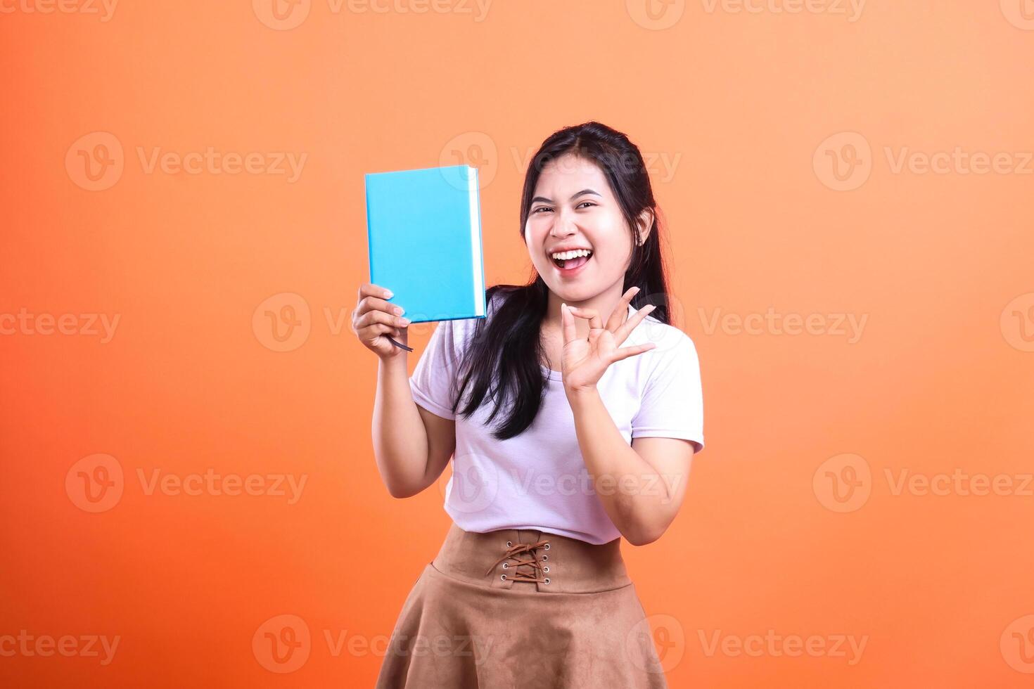 A smiling woman holds a blue book in one hand while gesturing with the other against a vibrant orange background photo