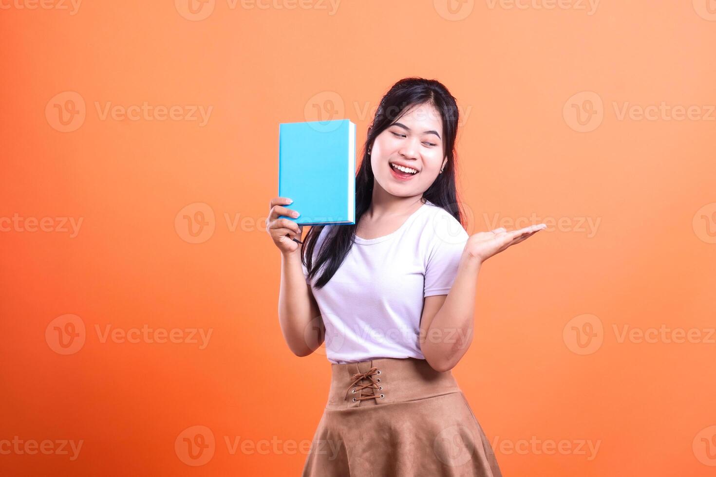 A young woman with long dark hair is holding a blue book while smiling broadly and gesturing with an open hand photo