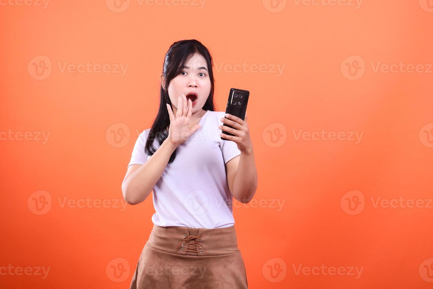 Woman with surprised expression holding smart phone isolated on orange background photo