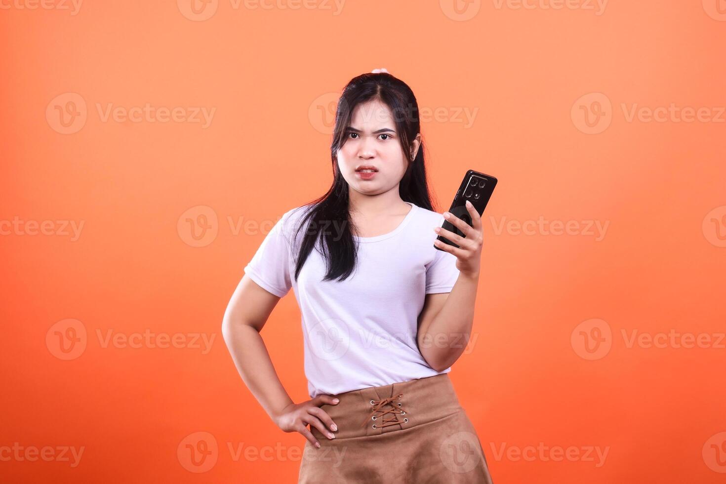 Woman in white shirt with smartphone on orange background poses with hands on hips photo