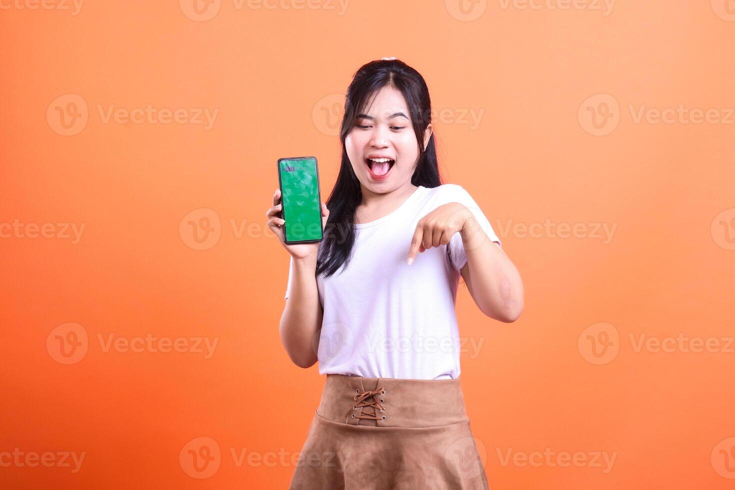 A woman asia holding a smart phone device with a green screen and pointing her index finger down with a happy expression and wearing a white shirt and brown skirt isolated on an orange background photo