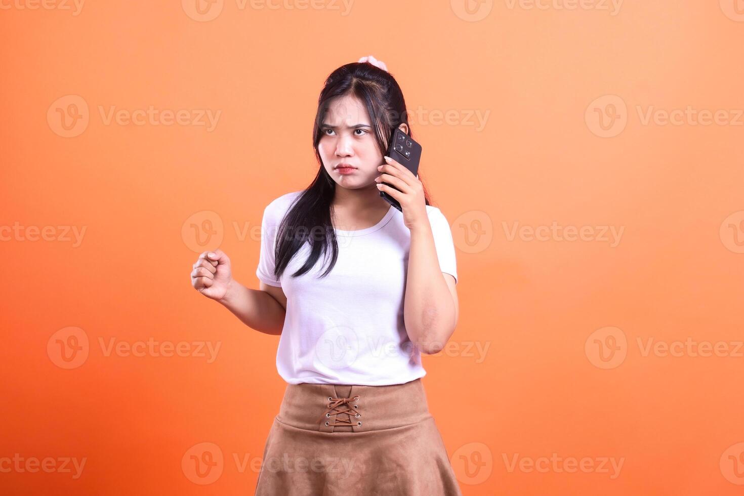 Young woman with long dark hair, wearing a white t-shirt and brown skirt, holding a cellphone on an orange background photo