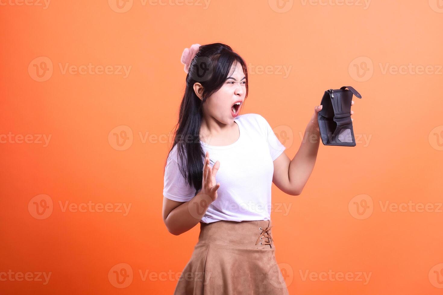 A young woman wearing a white shirt and brown skirt holding an empty black purse looking to the left and her mouth screaming in annoyance, isolated on an orange background photo