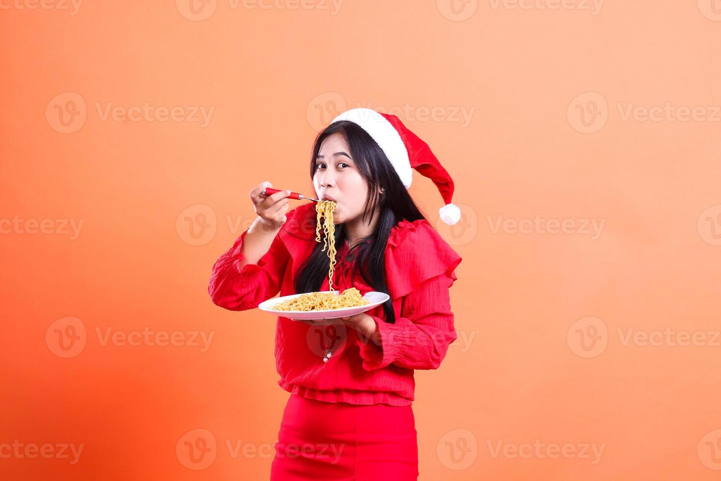 expression of girl wearing christmas dress, santa hat, right side view eating noodles front view, hand holding fork filled with noodles and white plate filled with noodles, isolated orange background photo