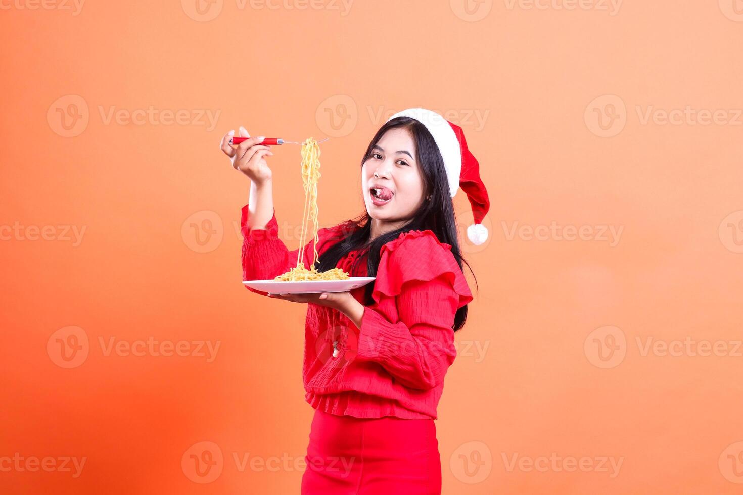 portrait of adult girl wearing christmas dress, santa hat, hungry looking at camera, hand holding fork filled with noodles upwards and white plate filled with noodles, isolated on orange background photo