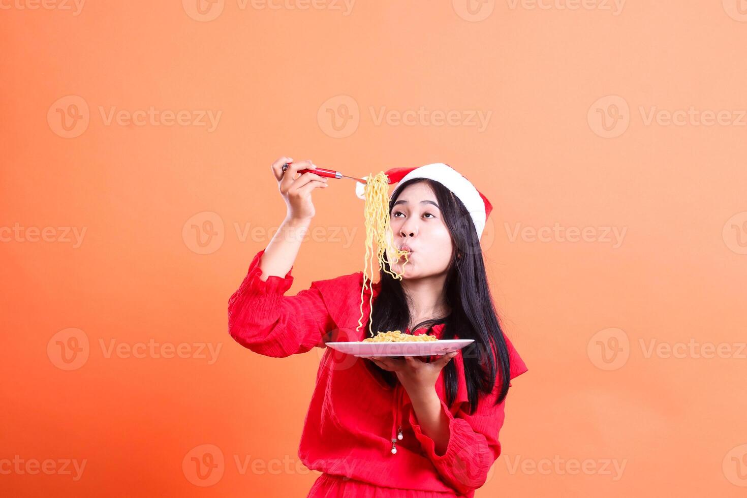 portrait of Asian female wearing Christmas blouse, Santa hat, enjoying slurping noodles, hand holding fork filled with noodles in front of head and white plate filled with noodles, isolated photo
