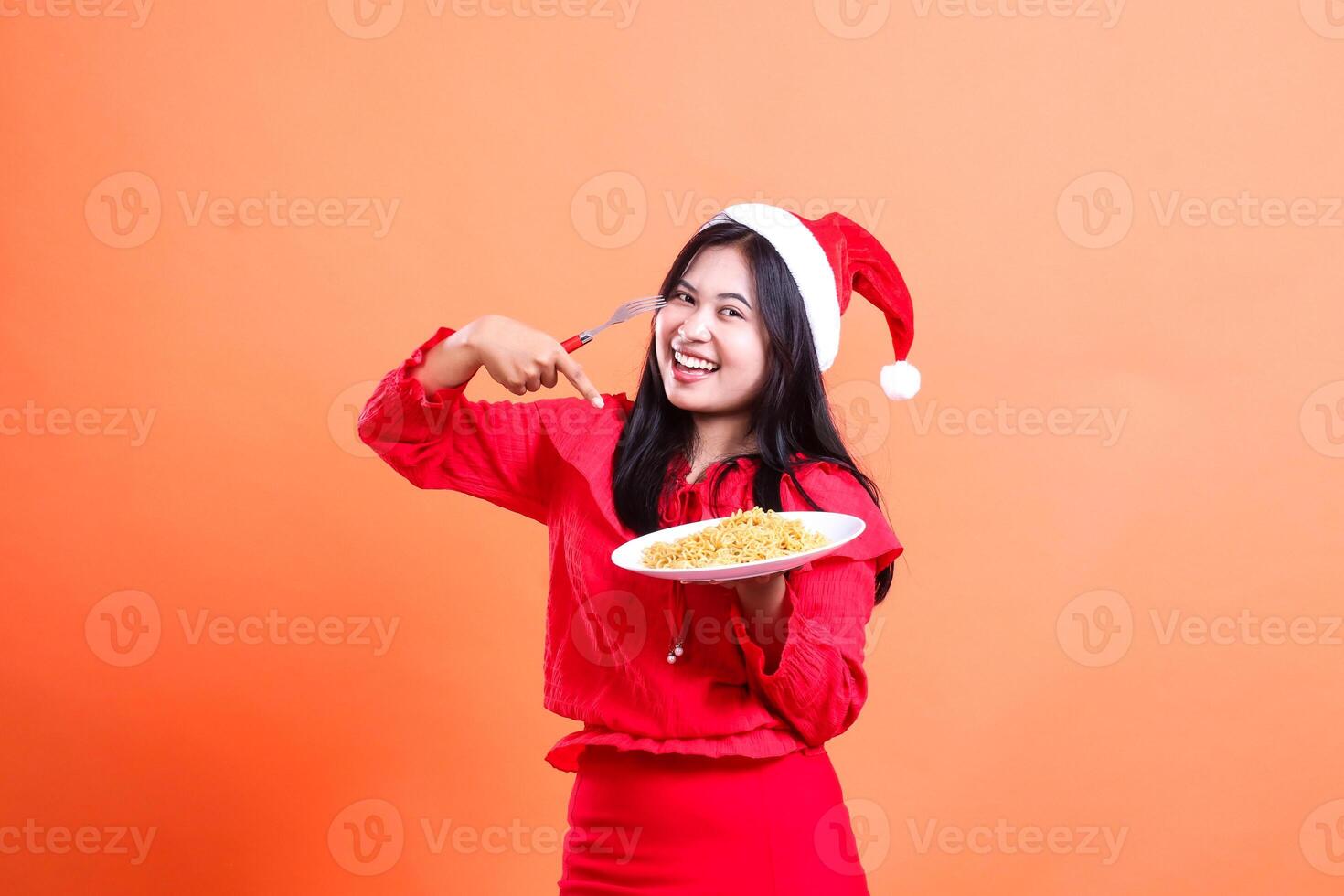 expression of adult woman wearing christmas dress, santa hat, cheerful looking at camera, hand holding fork and white plate filled with noodles while pointing at it, isolated on orange background photo