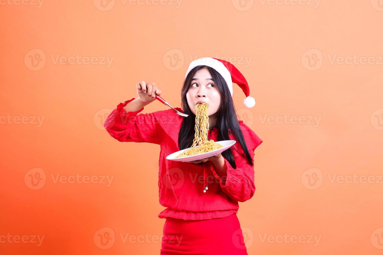 Asian ladies beauty surprised to feel noodles to the left, hand holding fork and white plate filled with noodles, wearing red christmas blouse, isolated on orange background photo