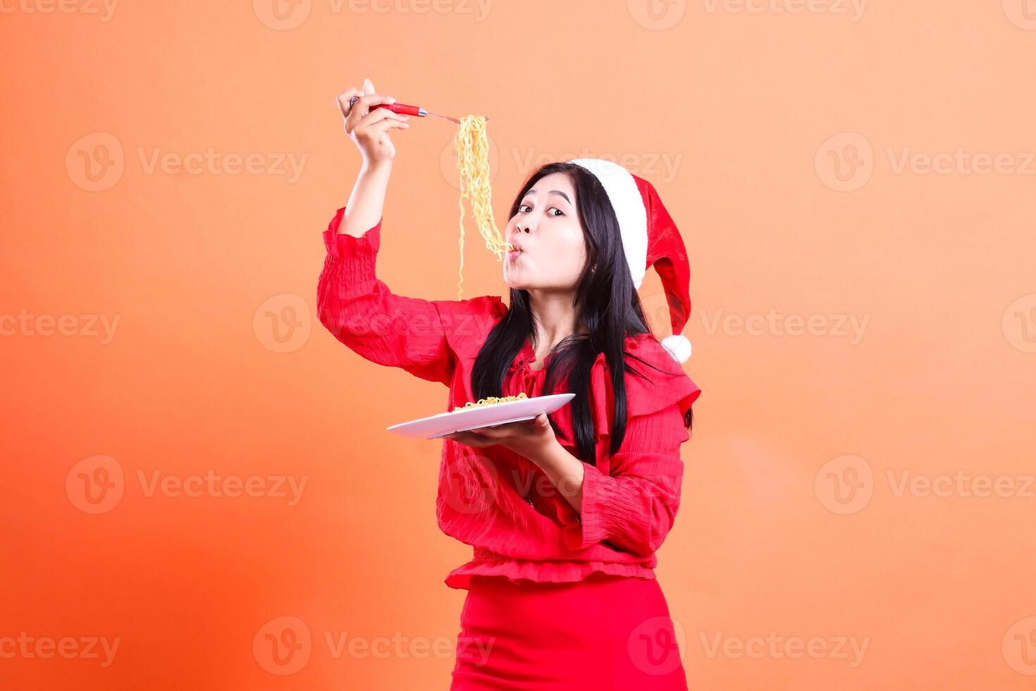 gesture of girl wearing red christmas blouse, Santa hat, left slurping noodles front view, hand holding noodle fork up and white plate filled with noodles, isolated on orange background photo