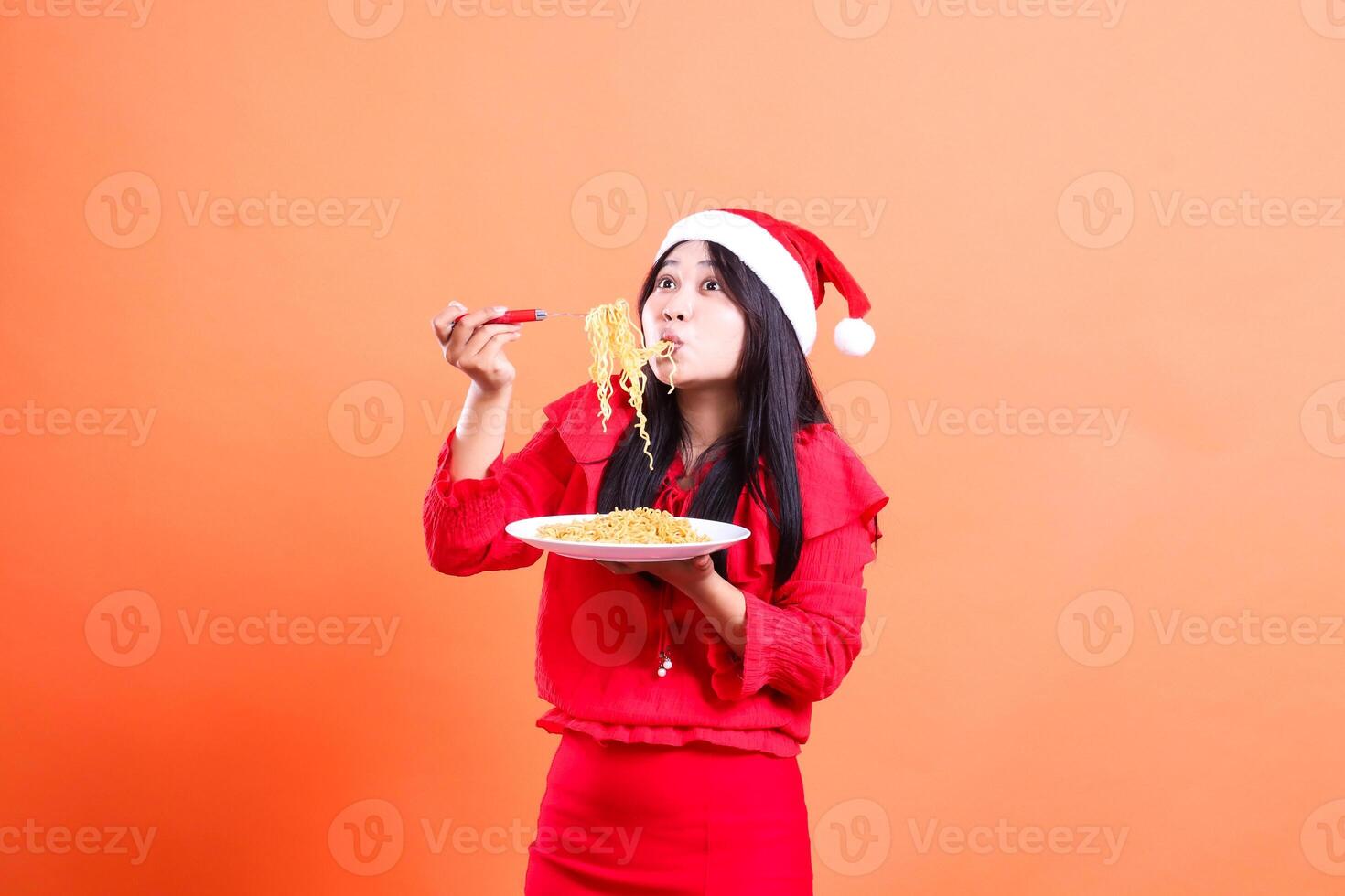 portrait of woman wearing christmas blouse, Santa hat, right side slurping noodles upward, hand holding noodle fork upward and white plate filled with noodles, isolated on orange background photo