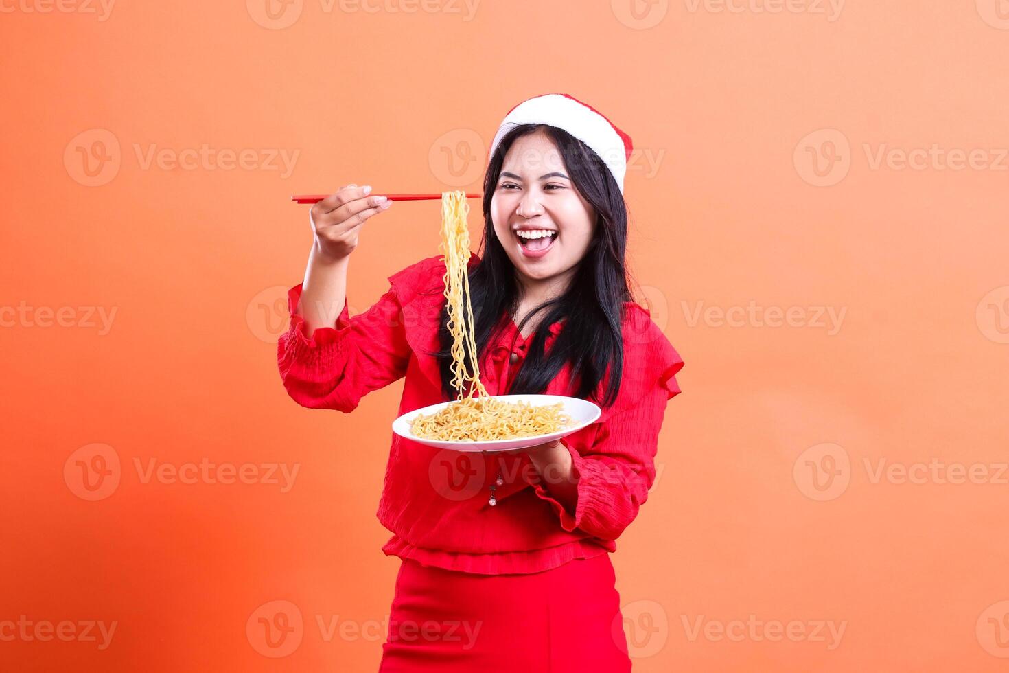 gesture of beautiful female wearing red christmas blouse, Santa hat, happy to noodles, hand holding chopsticks to mouth and white plate filled with noodles, isolated on orange background photo