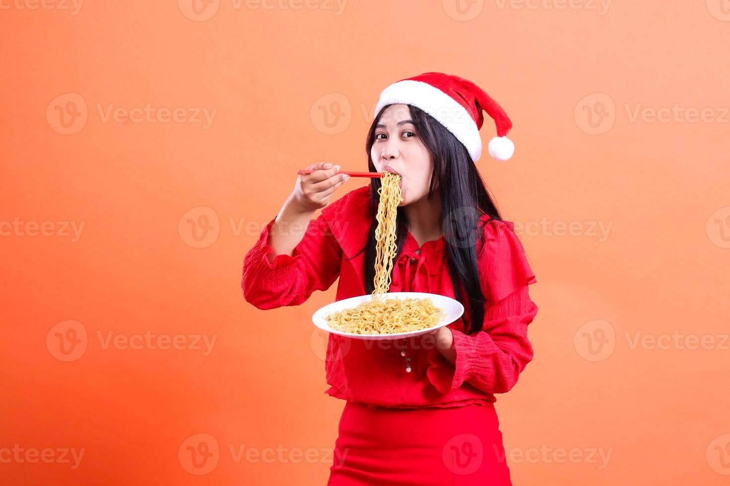 gesture of beautiful female wearing red christmas blouse, Santa hat, enjoying eating noodles to camera, hand holding chopsticks to mouth and white plate filled with noodles, isolated orange background photo