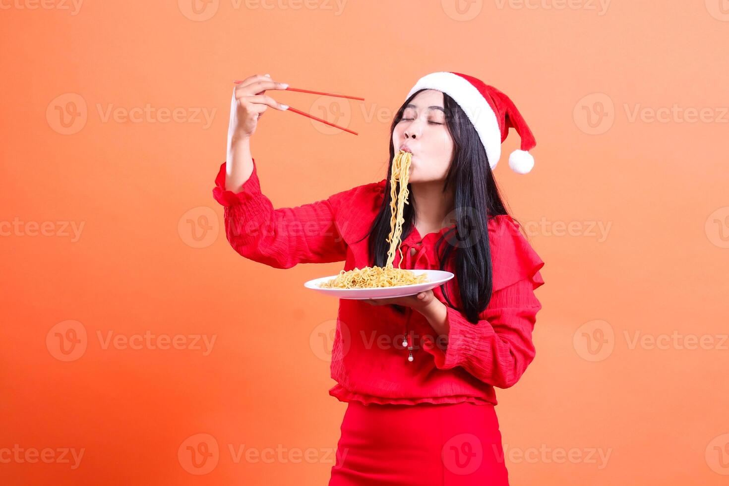 expression of asia woman wearing christmas dress, santa hat, slurping noodles with closed eyes, hands holding chopsticks and white plate filled with noodles, isolated on orange background photo