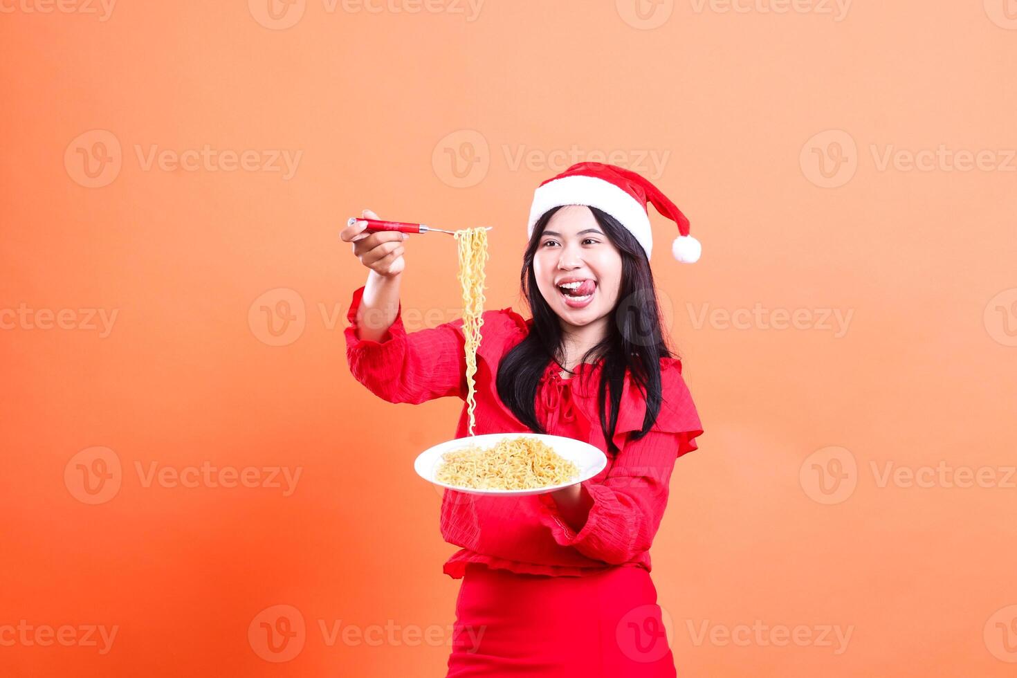 expression of young women wearing christmas dress, santa hat, enjoying looking at noodles, hand holding noodle fork up and white plate filled with noodles in front, isolated on orange background photo