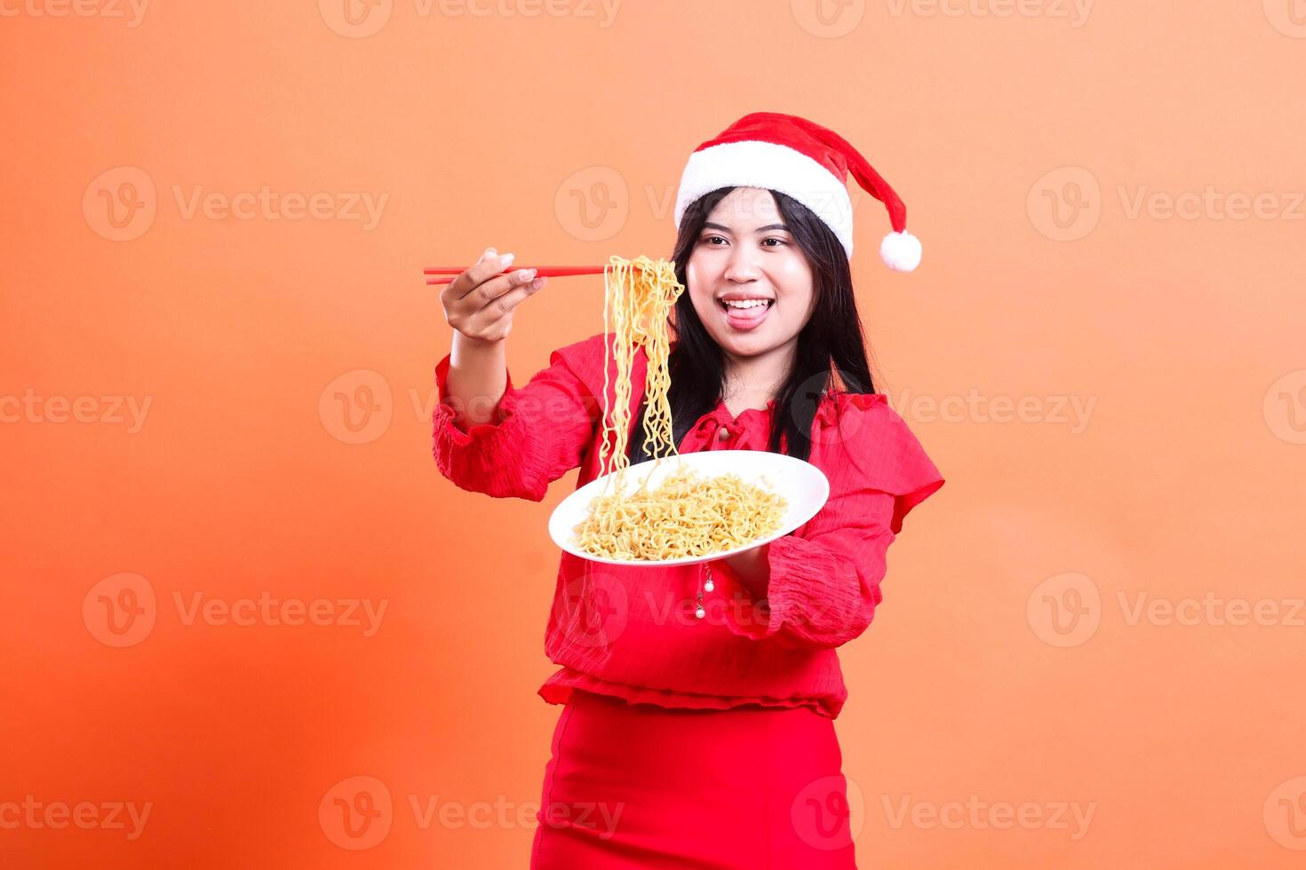 Asian adult ladies happy eating noodles, hand holding chopsticks filled with noodles up and white plate filled with noodles forward, wearing red Christmas blouse, Santa hat, isolated orange background photo