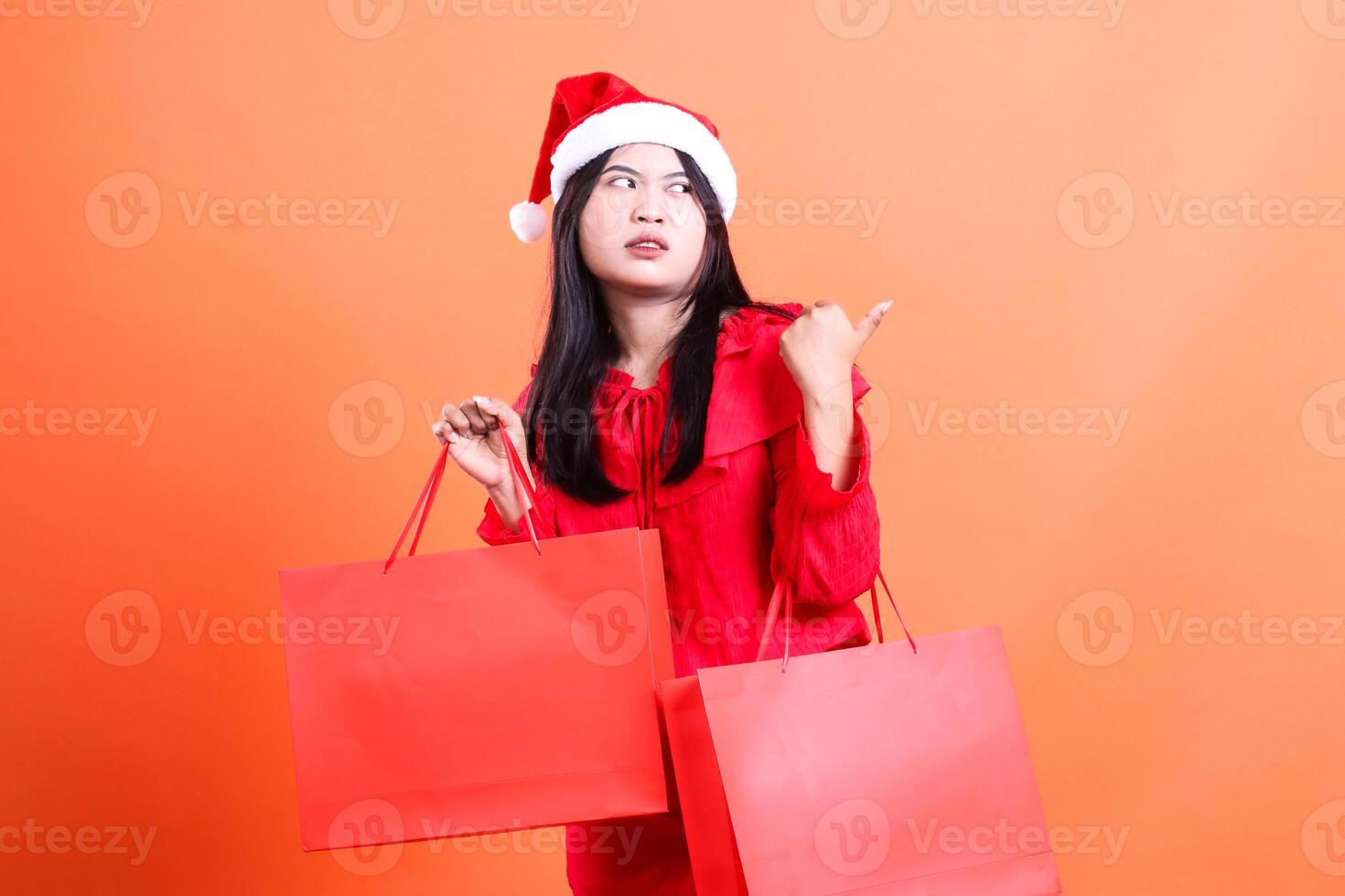 Asian adult woman expression wearing red blouse and santa hat, standing disgusted looking to the left hand holding red bag of christmas gifts and pointing left, isolated on orange background photo
