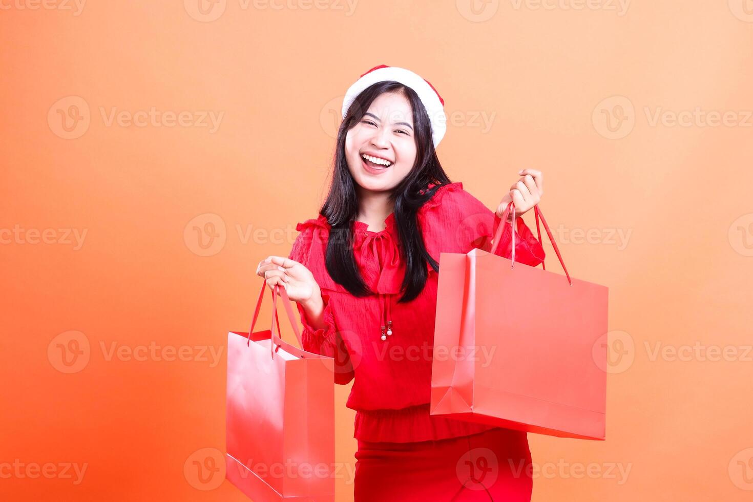 young woman expression wearing red blouse and santa hat, standing happily in front of both hands lifting red bag of christmas gifts, isolated on orange background photo