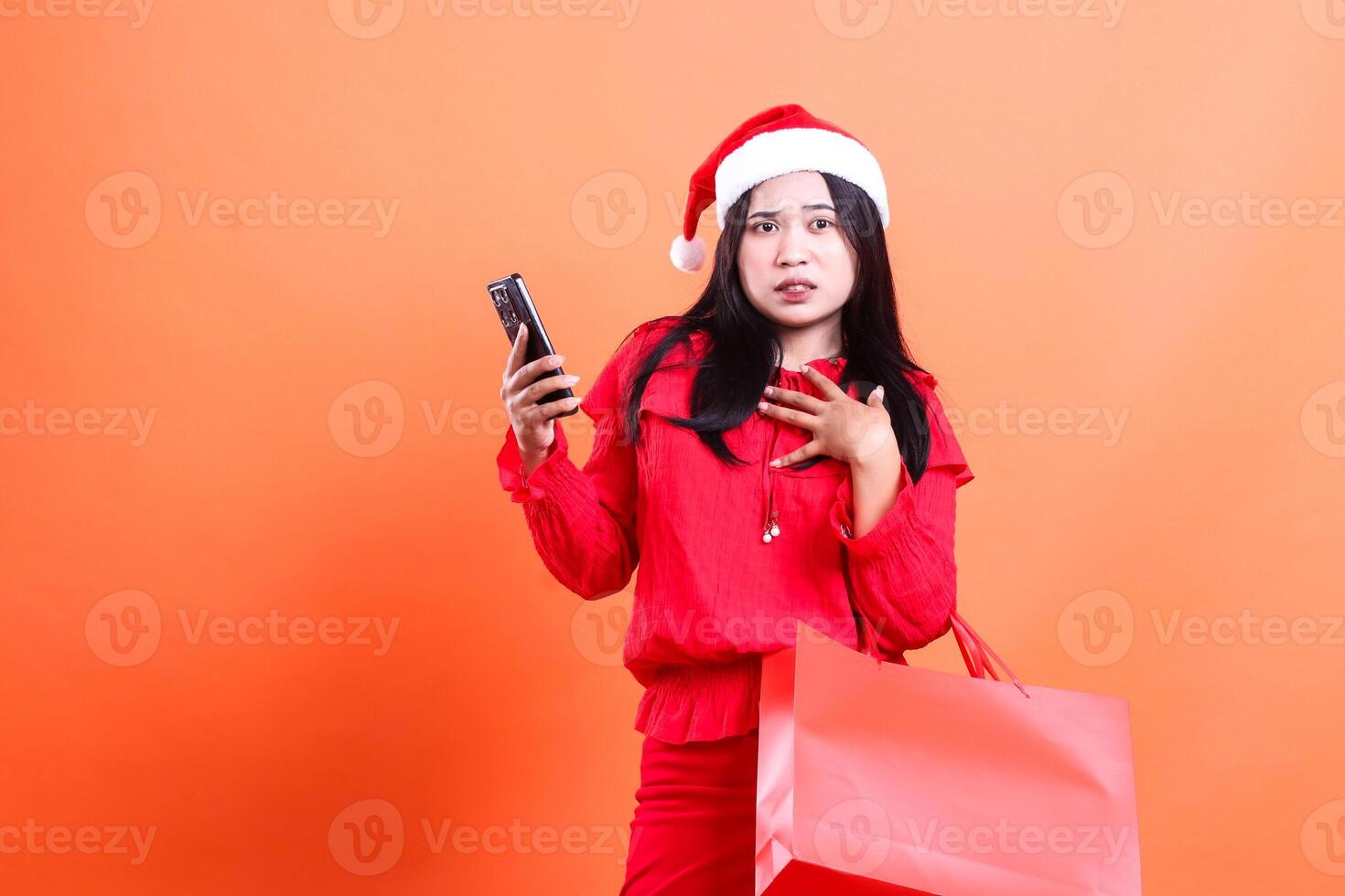 expression of ladies wearing christmas dress, santa hat, disgusted afraid looking at camera, hands dumbfounded using gadget, carrying red bag of christmas gifts, isolated on orange background photo