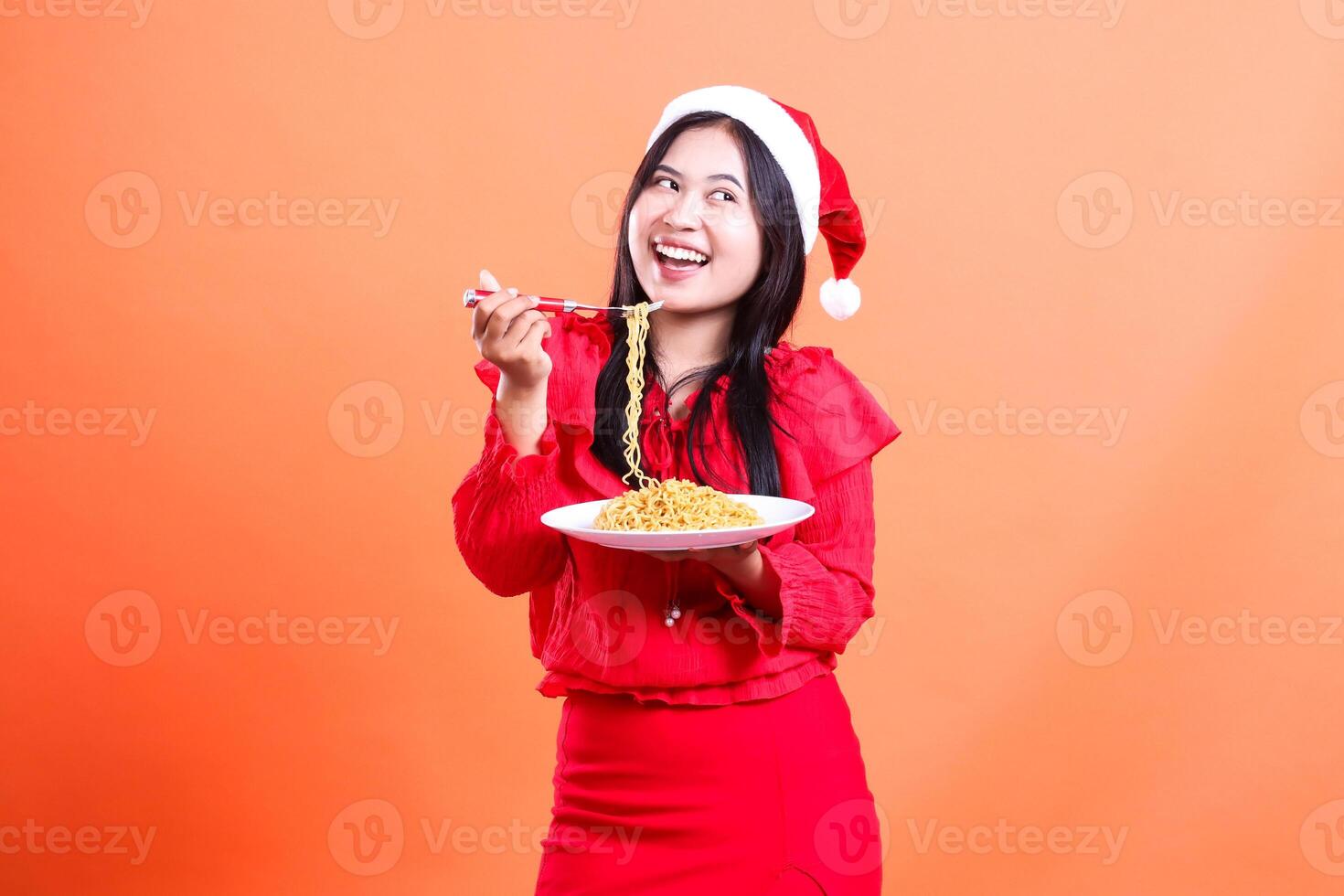 gesture of beautiful female wearing red christmas blouse, Santa hat, cheerful to left tilted, hand holding fork filled with noodles from white plate commemorating christmas ,isolated orange background photo