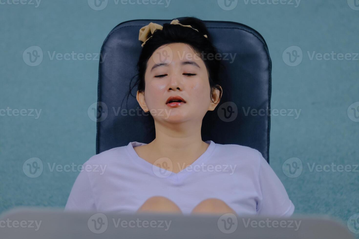 A woman is shown from the chest up, sitting and resting on a black fitness machine. Her eyes are closed and her mouth is slightly open. photo
