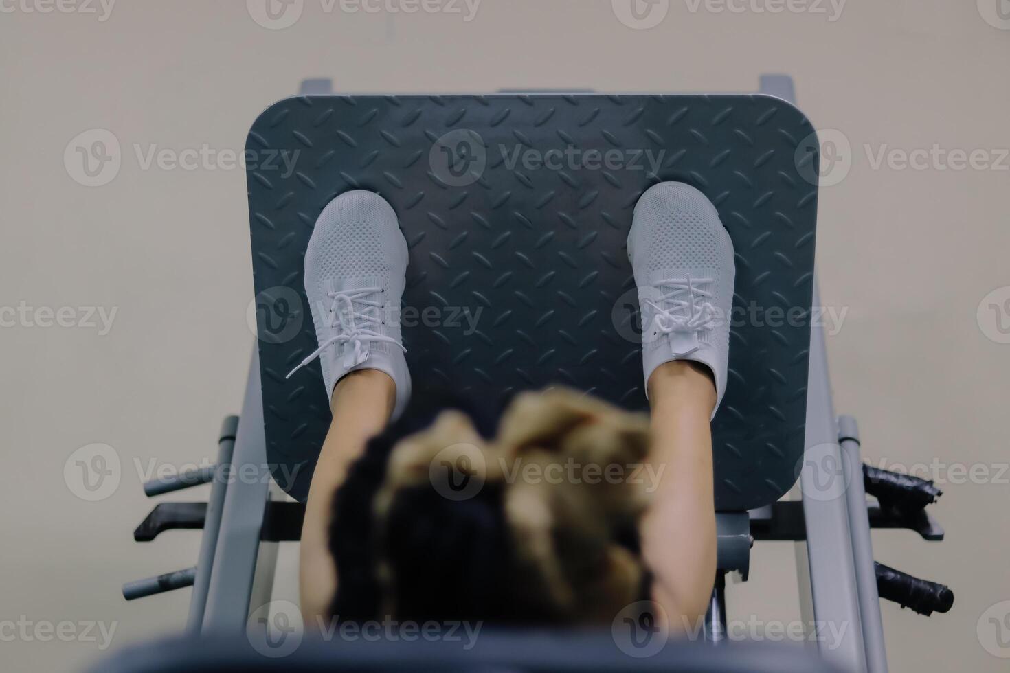 close up of a woman's head using a leg press machine in a fitness center environment. Her feet are wearing white shoes, positioned on a gray metal platform with a textured surface. photo