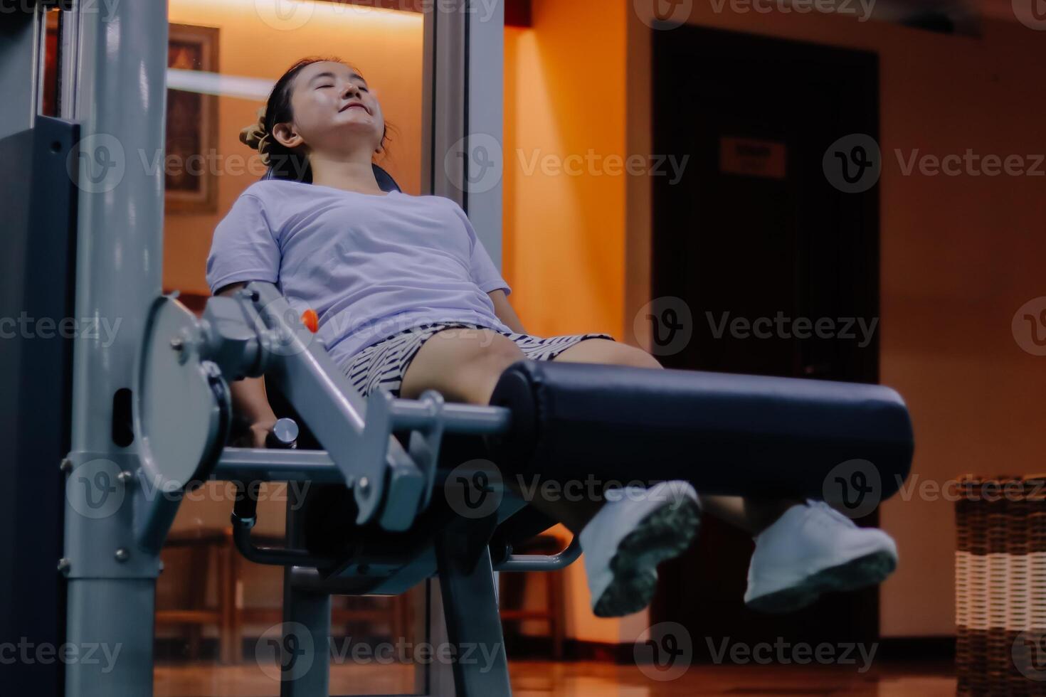 woman wearing purple shirt, shorts, and white shoes, is using a leg extension exercise machine. The person's face is visible, and her eyes are closed. The scene takes place indoors. photo