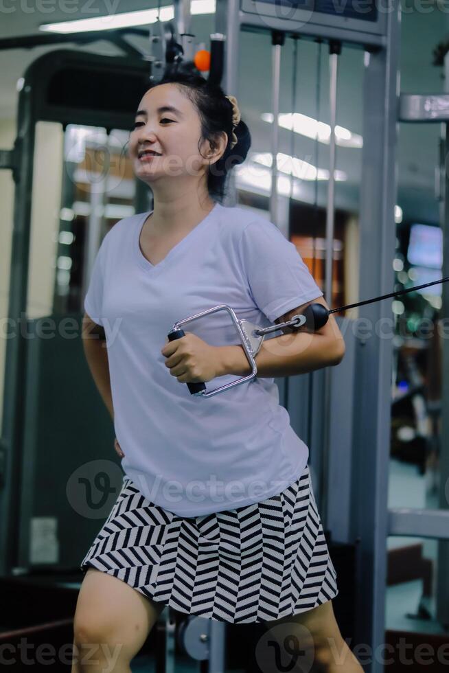 A woman at an indoor gym, actively engaged in an exercise routine using a cable machine. She smiles as she holds the handles attached to the cable. photo