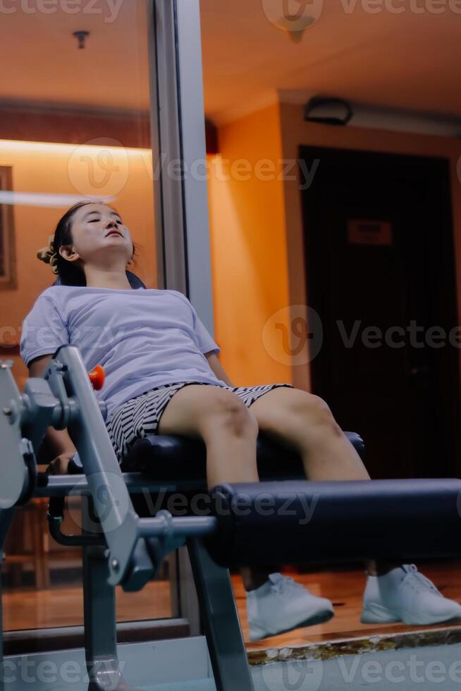 A woman uses a leg extension machine in a fitness center. She sits with her legs on the bar of the machine. The background is a window with lights, a door and some equipment. photo