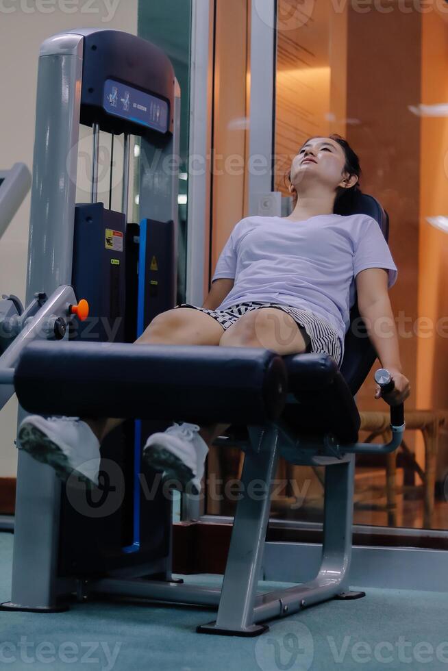 woman using leg extension machine at gym. sitting with legs positioned under padded machine bar. wearing white shirt, short skirt, and white shoes photo