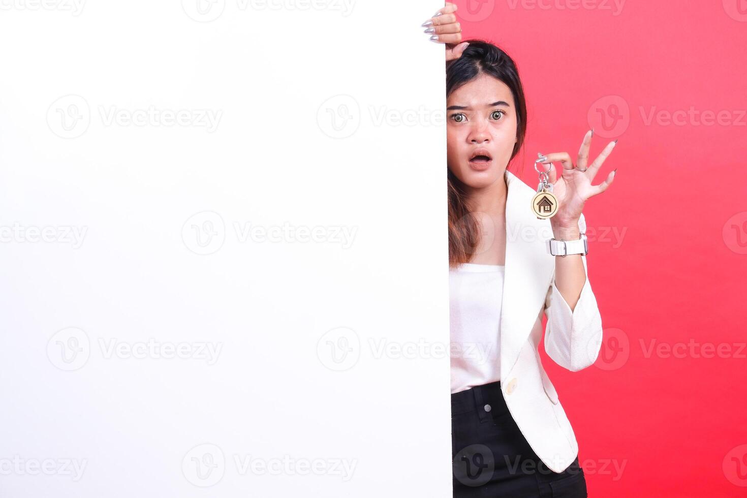 A woman is holding a house key and hiding behind a white banner with a surprised expression, isolated on a red background photo