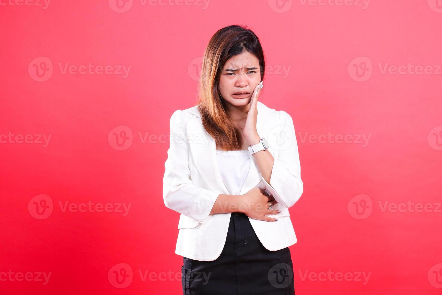 A woman with light brown hair wearing a white jacket and black skirt is touching her cheek, seemingly having a toothache, isolated on a red background photo