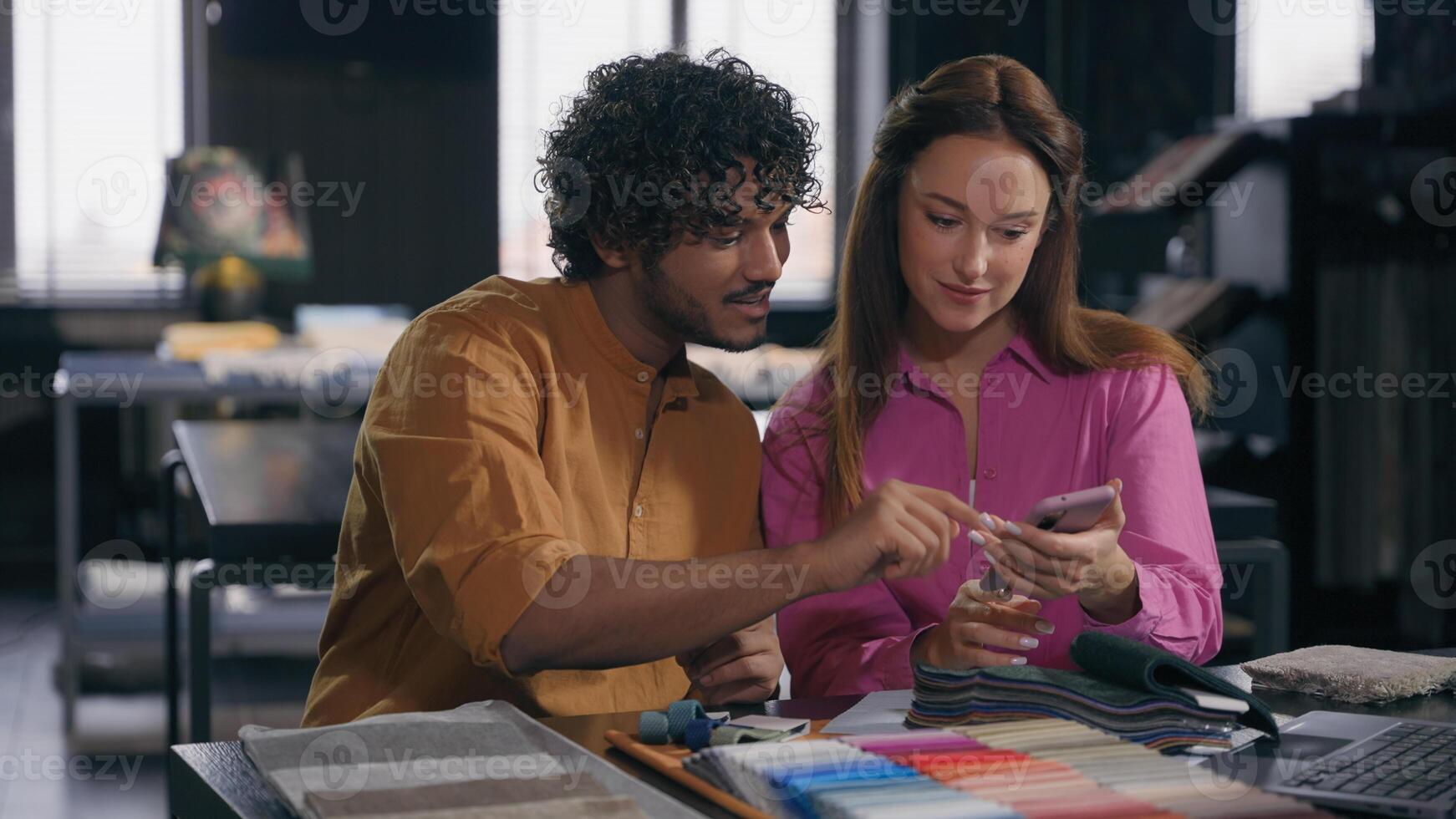 Multiracial couple creative designers work in modern office studio Arabian Indian man Caucasian woman using phone choosing perfect sample of material color tone for new house interior design project photo