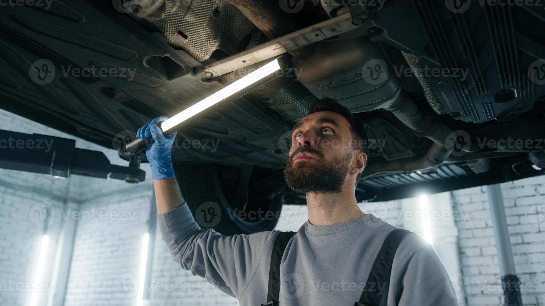Caucasian man professional mechanic in uniform repairman male engineer working underneath car check fix problem under automobile using flashlight light vehicle diagnostics garage service auto repair photo