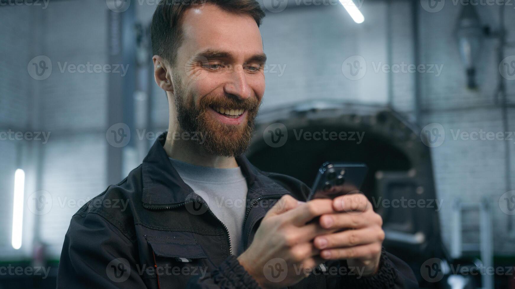 Auto mechanic man Caucasian male manager worker maintenance engineer looking at mobile phone screen cheerful laughing smartphone using in car repair service station on open automobile hood background photo