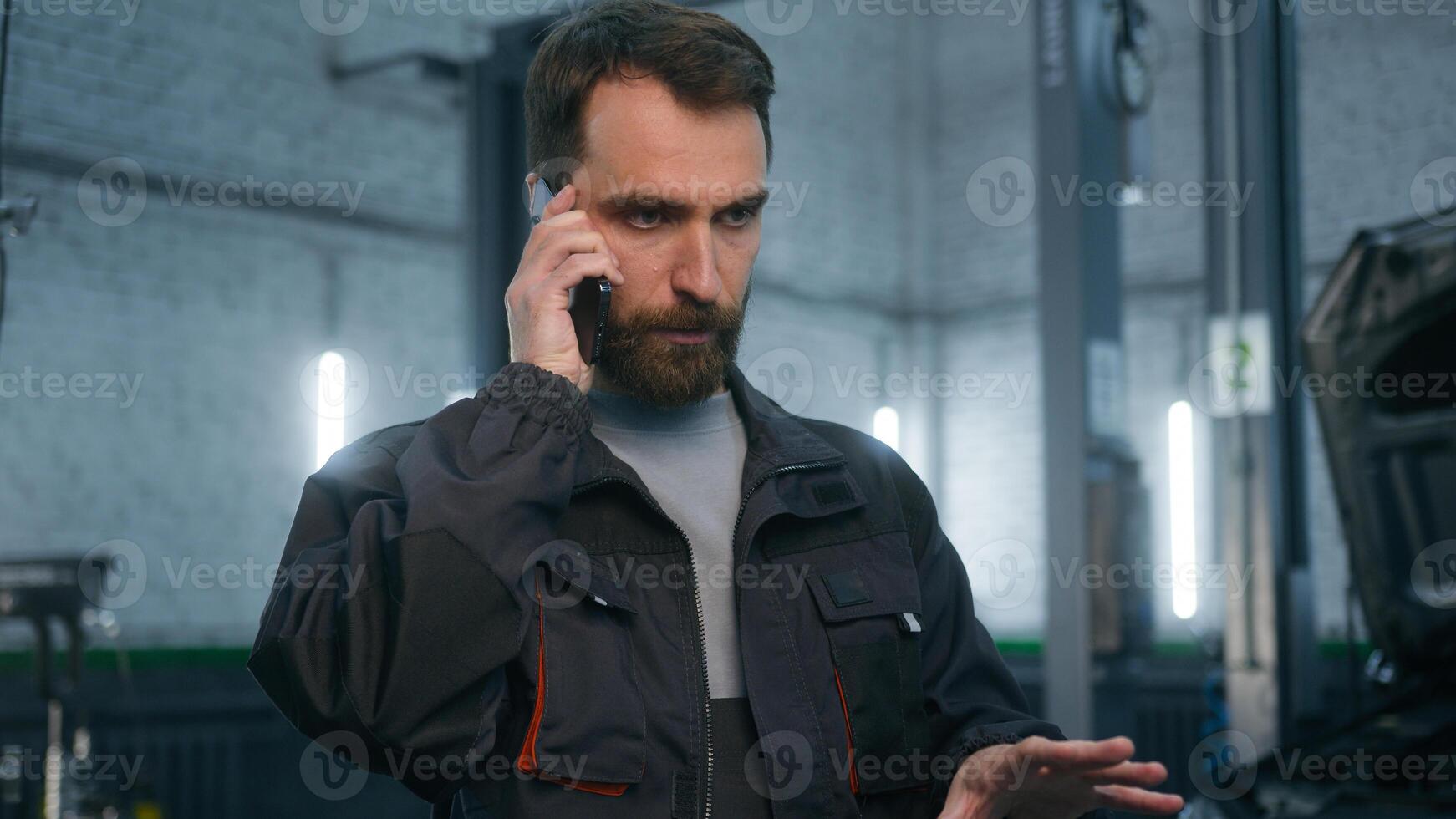 Caucasian man professional worker auto engineer mechanic car repairman working in automobile service station bearded male guy talking phone smartphone in garage answer mobile call speaking with client photo