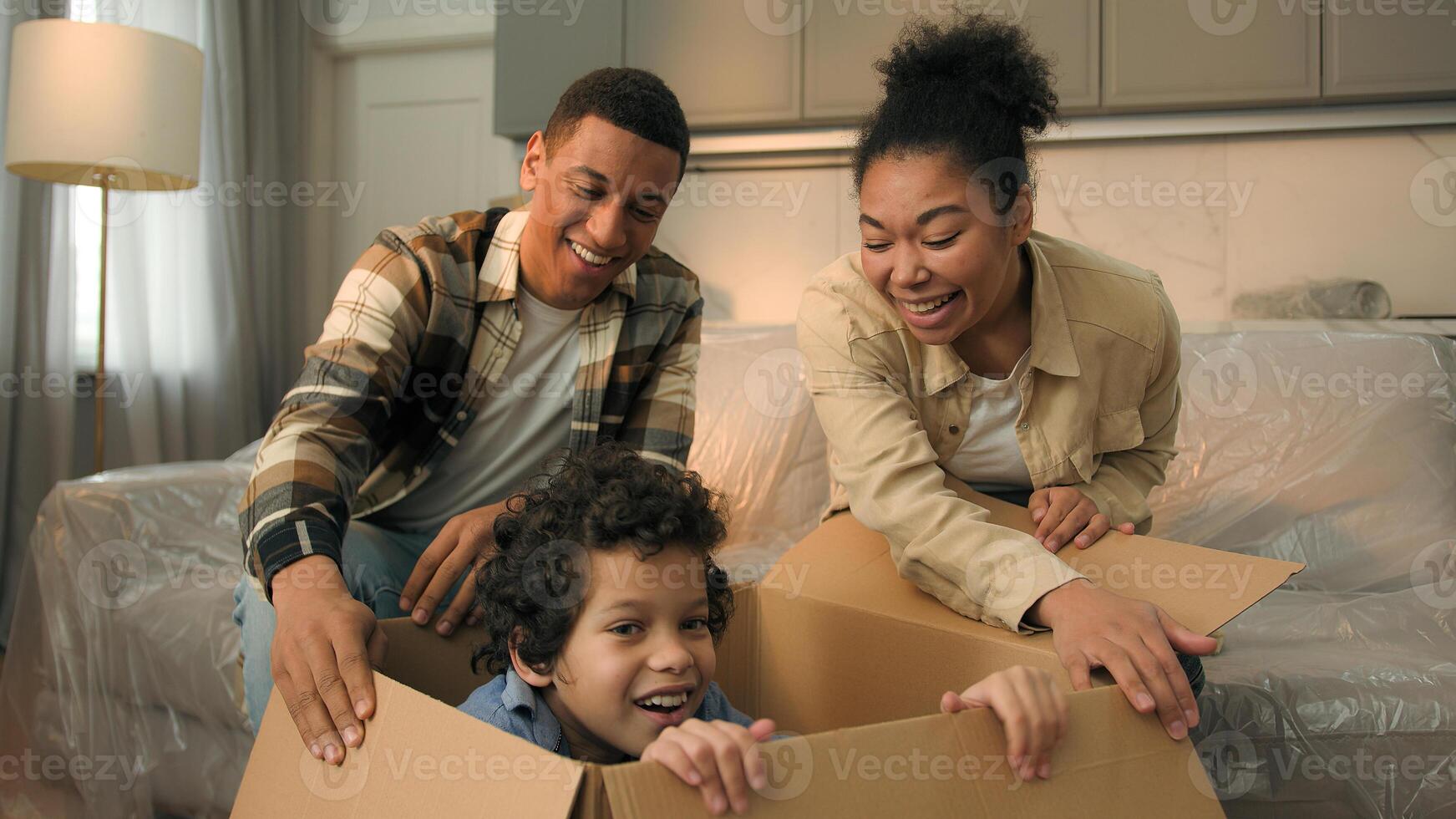 African American family moving in new home relocation happy parents unpacking parcel surprise funny joyful son little boy child jumping out of cardboard box playing game hide and seek kid having fun photo