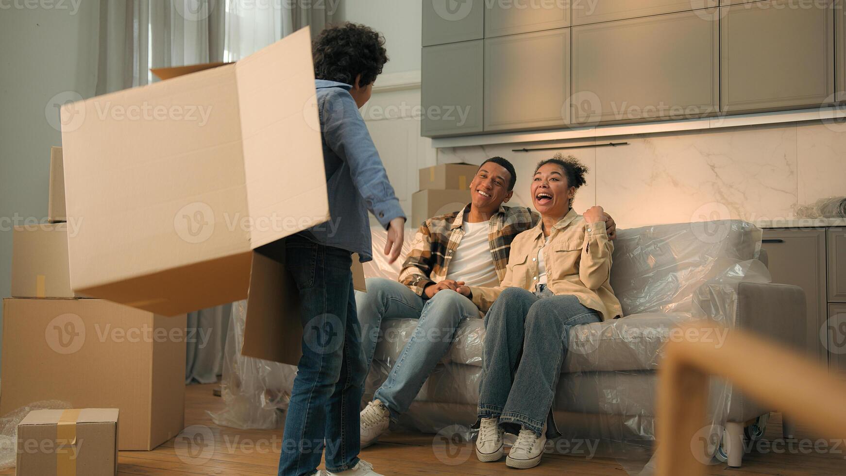 African American family mom dad on sofa home happy naughty little son child playing hide and seek game funny kid jumping out from box play with parents having fun unpacking boxes relocation moving day photo