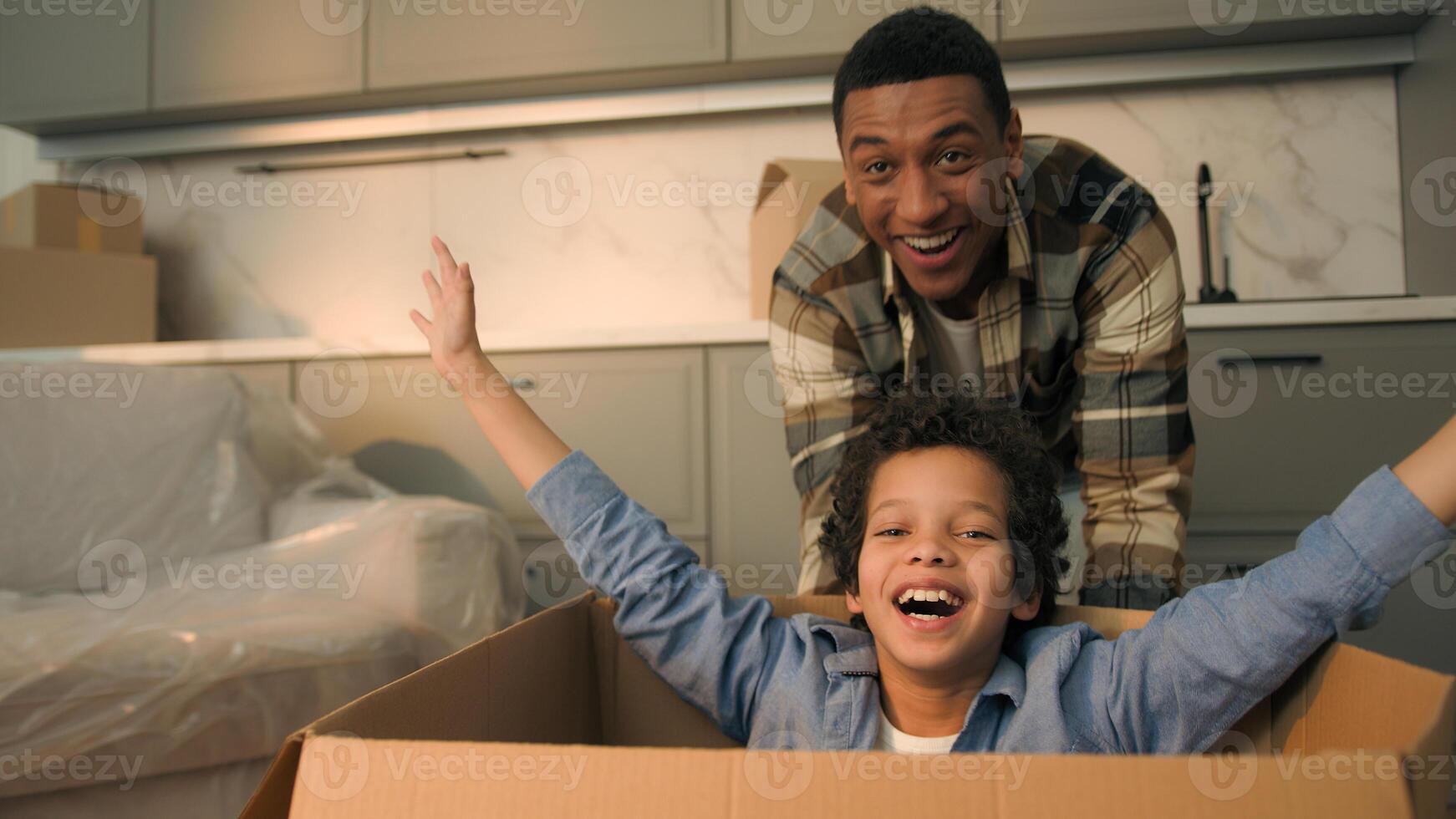 Happy African American family having fun celebrate relocation new home father run push cardboard box with little son boy child inside joyful smiling kid dad playing together moving day credit mortgage photo