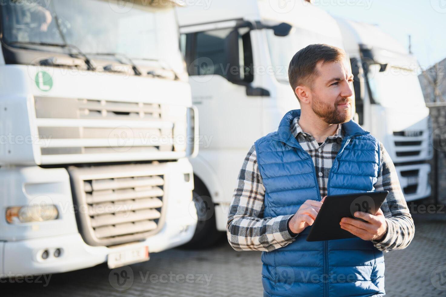 Handsome Caucasian man wearing red cap standing next to trucks while using tablet device. Hardworking driver typing message or text for delivery order Logistics company concept photo