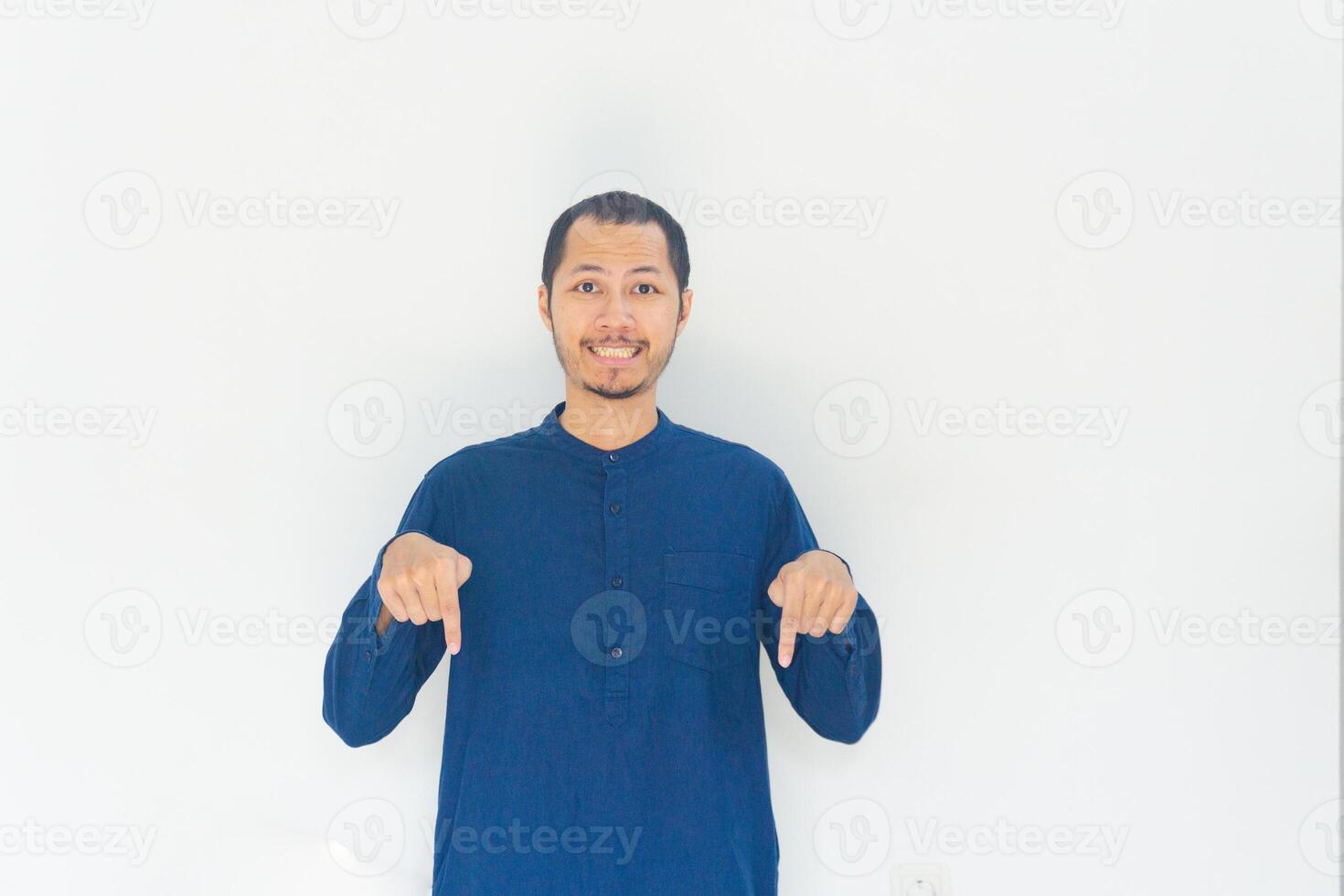 Muslim men wearing blue baju koko is pointing down and smiling in isolated white background photo