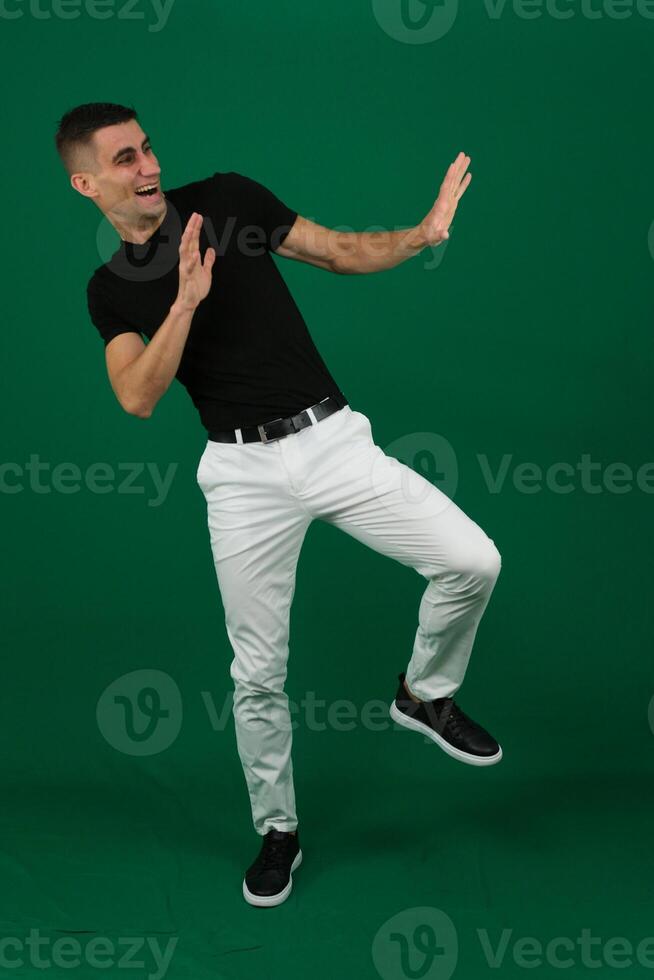 Young man celebrating success. Happy funny joyful excited guy in stylish green party suit and cool glasses raising fist up and dancing isolated on bright yellow background. Full length shot, side view photo